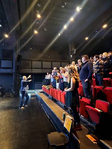 Audience members standing in tiered theatre seating, following guidance from a conductor standing at the front of the auditorium.