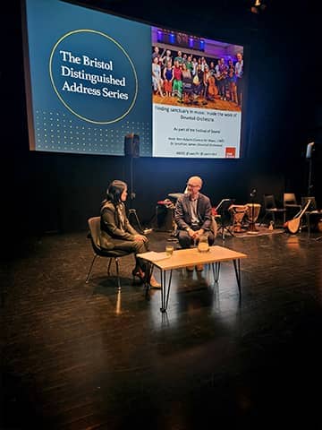 Two speakers seated on stage in conversation, with musical instruments and event branding visible in the background.