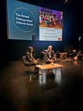 Two speakers seated on stage in conversation, with musical instruments and event branding visible in the background.