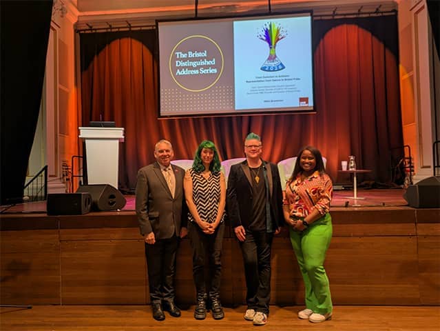 Photo of four event speakers standing together in front of the stage, with the Bristol Distinguished Address Series banner projected above them.