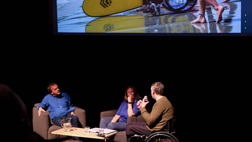 Two white men, one in a wheelchair, and a woman in chairs on a stage beneath a screen with surfers on it.