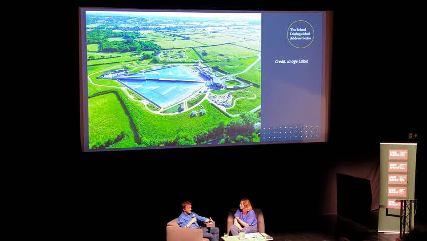 A white man and a white woman in chairs on a stage beneath a large screen with an image of green fields and a large blue swimming pool in the centre.
