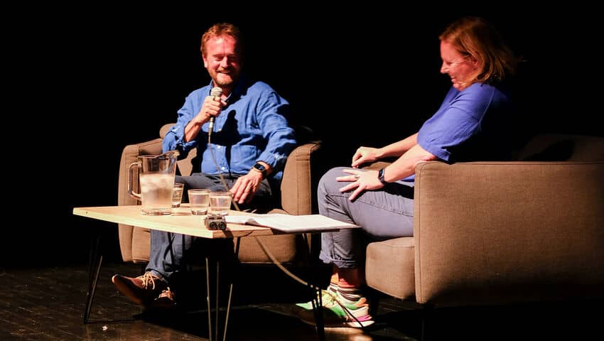 A white man sits in a chair holding a microphone opposite a white woman