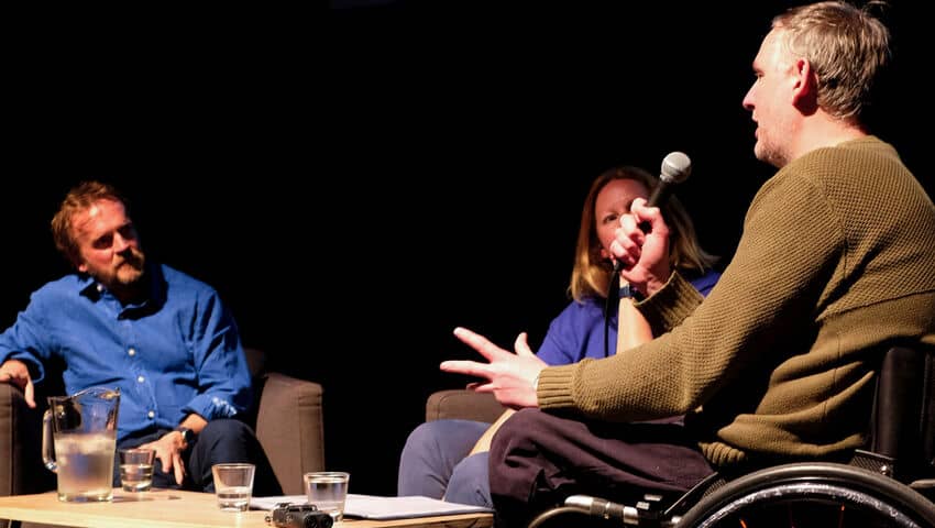 Two white men, one in a wheelchair, and a woman in chairs on a stage having a public discussion.