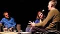 Two white men, one in a wheelchair, and a woman in chairs on a stage having a public discussion.