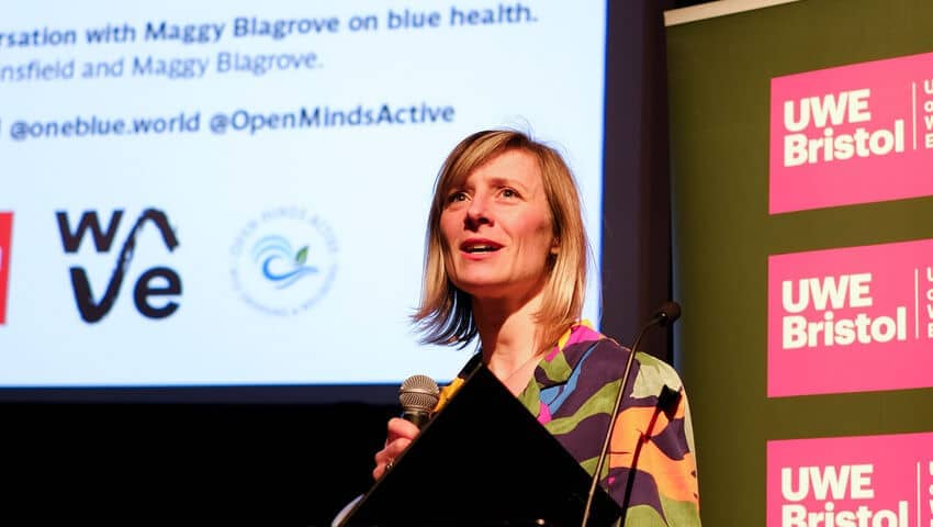 A white woman at a lectern with a UWE Bristol banner behind here and the WAVE logo on a screen also behind her.