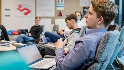 Student sat back in a classroom with a laptop on his knees with other students in the background.