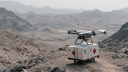 Image of a drone carrying a white container with the medical red cross logo on it against a background of a dry environment.