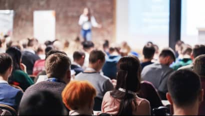 A woman presenting beside a poster to a busy conference hall filled with people.