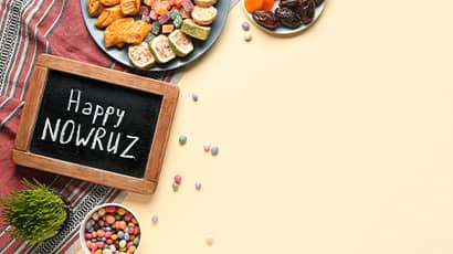 A picture featuring traditional Nowruz food and snacks on a light yellow background and a small blackboard with 'Happy Nowruz'.