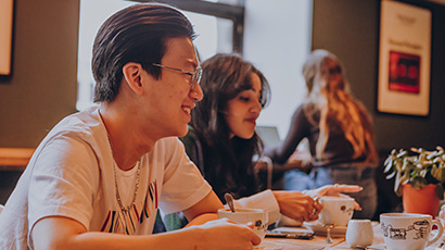 Two students at a table with coffee cups talking in a cosy coffee shop setting.