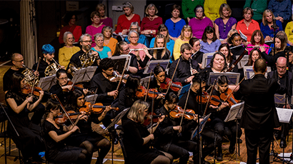 Large group of singers in colourful tops and orchestral players