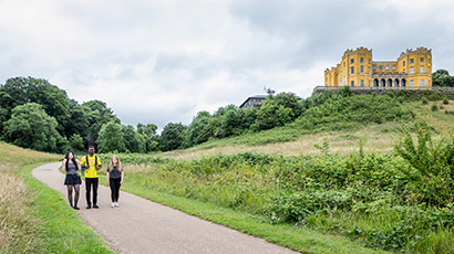 Three people walking along a path in Stoke Park with the yellow mansion on the hill in the background. 