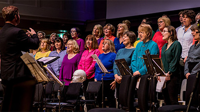 A choir in colourful tops performing on stage with a conductor guiding them.