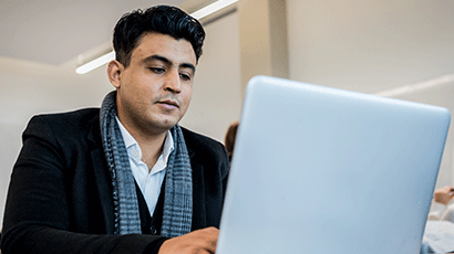 Man in smart jacket and scarf working at his laptop in a classroom.