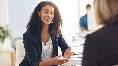 A woman in smart business dress talking to another colleague across a desk.