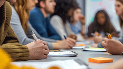 A blurred image of a group of people sat around a large desk making notes and looking up at a presenter off camera.