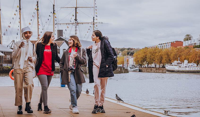A group of students walking alongside the waterfront in Bristol talking with the SS Great Britain in the background.