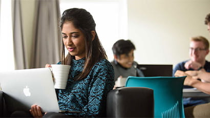 Student sitting in common room on laptop holding a mug of coffee.