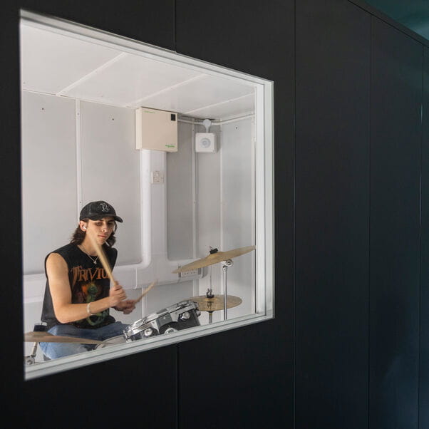 A white man through a window of a steel container in a band T-shirt wearing a cap drumming.