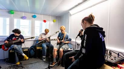 Three students with guitars sitting down and one with a microphone sitting on a table in a pracitice space.