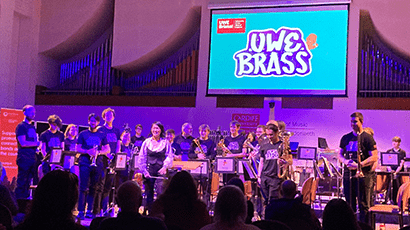 brass band standing on stage with large UWE Brass blue logo on the screen