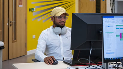 A bearded Asian male wearing a baseball cap seated in front of a computer in Launch Space.