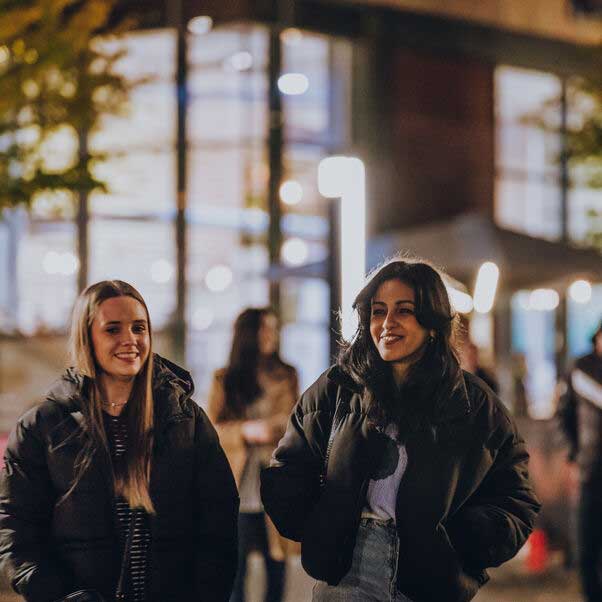 Couple of smiling girls walking together in the evening with the light of a building behind them. 