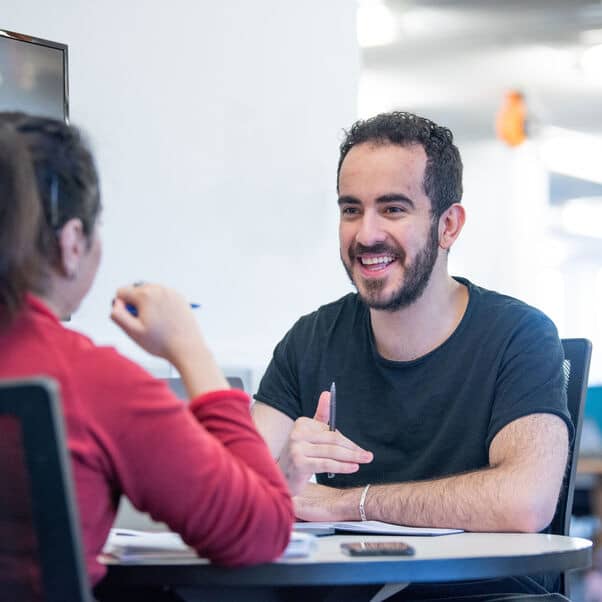 A staff member sat smiling behind a desk talking to a student