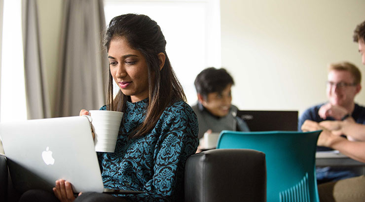 Students in a living room; one of them working on a laptop