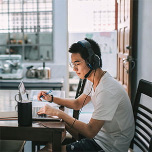 Male student sitting in a cafe while studying on their laptop and wearing headphones.