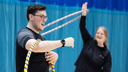 A student grapples with a hula hoop around his arm while friend looks on laughing and whirling a hoop around her hand during a MOVE class at the Centre for Sport. 