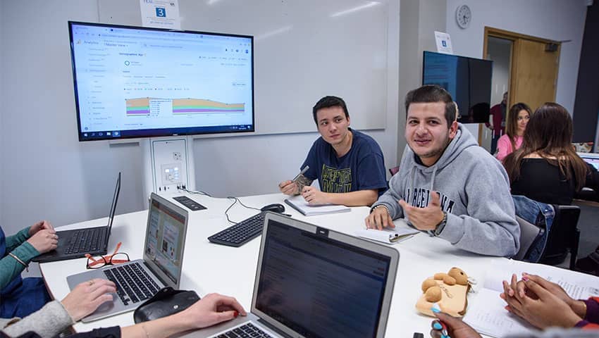 A group of students sat around a table with laptops with a monitor on the wall in front showing graphs.