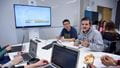 A group of students sat around a table with laptops with a monitor on the wall in front showing graphs.