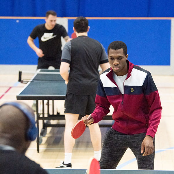 A man focuses intently on his MOVE table tennis game in the sports hall at UWE Bristol while another match takes place in the background.