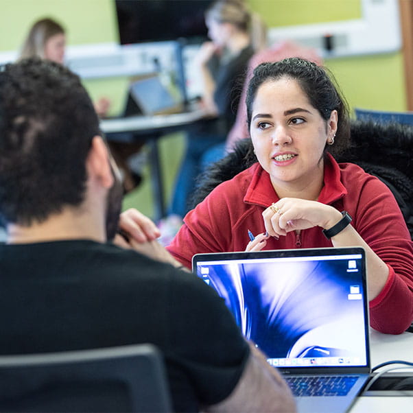 A student in a red jumper talks to another student, whose back is to camera, over their laptops.