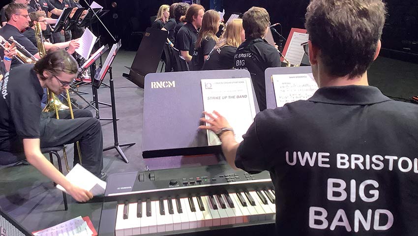 Young person seated at piano in front of other musicians with UWE Bristol Big Band polo shirt