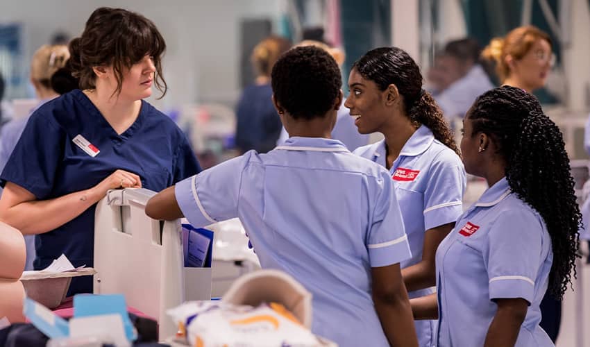 A group of nurses stood in a clinical setting in conversation with each other. The room is busy with various people working behind them. 
