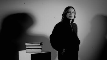 Black-and-white photograph showing a person standing in front of a plain wall with strong shadows cast on both sides. To the left, there is a small pedestal or table with a stack of books placed on top.