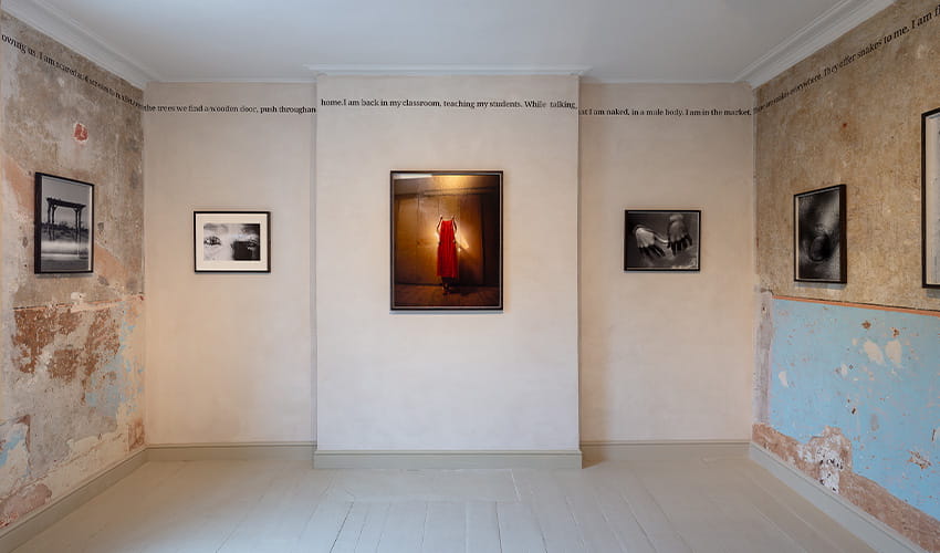 Art gallery room with worn, partially exposed plaster walls and a light wooden floor. Five framed photographs are displayed: two on the left wall, two on the right wall, and one centered on the back wall showing a vivid red dress. Text runs along the top edges of the walls.