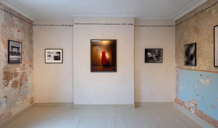 Art gallery room with worn, partially exposed plaster walls and a light wooden floor. Five framed photographs are displayed: two on the left wall, two on the right wall, and one centered on the back wall showing a vivid red dress. Text runs along the top edges of the walls.