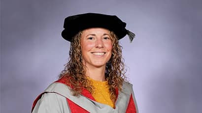 A woman wearing a university graduation mortar board and gown smiles against a grey background.