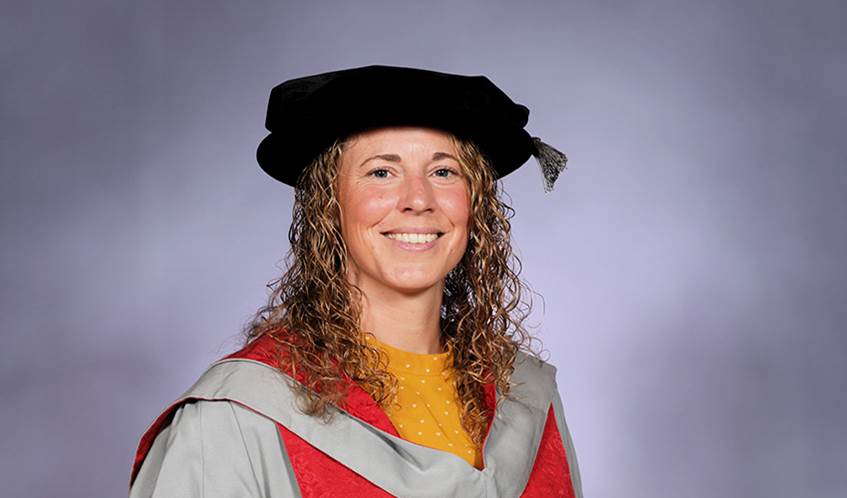 A woman wearing a university graduation mortar board and gown smiles against a grey background.