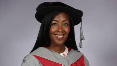 A lady smiles at the camera wearing a graduation cap and gown
