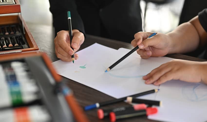 Two children sit at a table drawing with coloured pencils., while art supplies and an open box of pastels are visible alongside.