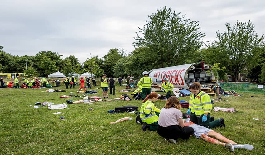 The image shows a major aviation disaster simulation in a field, with a shell of an aircraft in the background and lots of emergency service workers dealing with patients in the foreground.