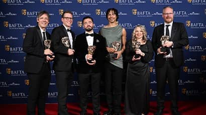 A group of six people smile at the camera holding BAFTA awards. 