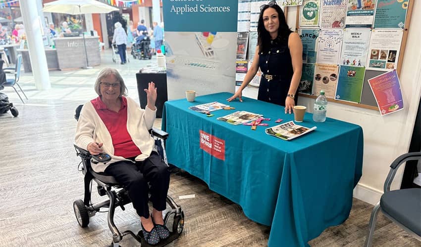 A person using a mobility scooter sits beside a UWE Bristol information table, while another person stands behind the table displaying leaflets and promotional materials.
