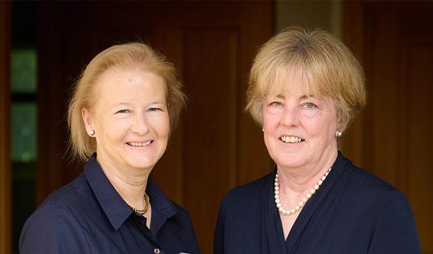 A headshot of two women smiling against a brown background