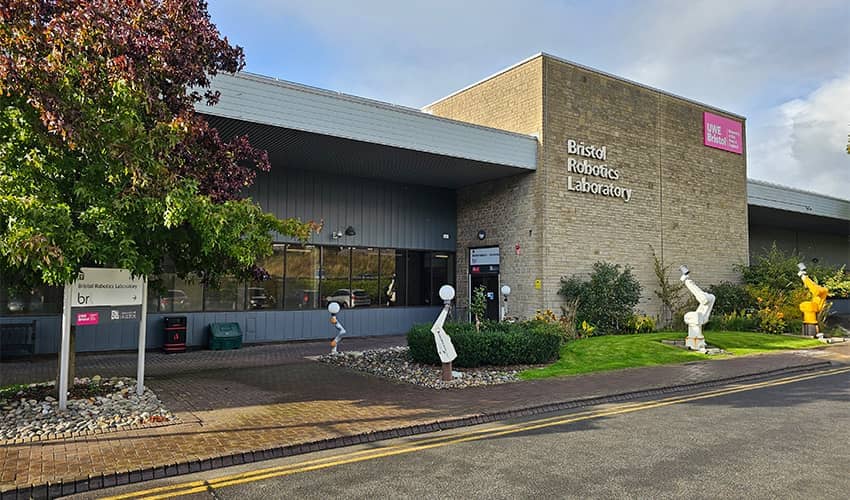 The exterior of a building which has a brick facade and robotic arm figures placed in front of the entrance to the building. The building has 'Bristol Robotics Laboratory' written on the outside. There are trees around the building. 